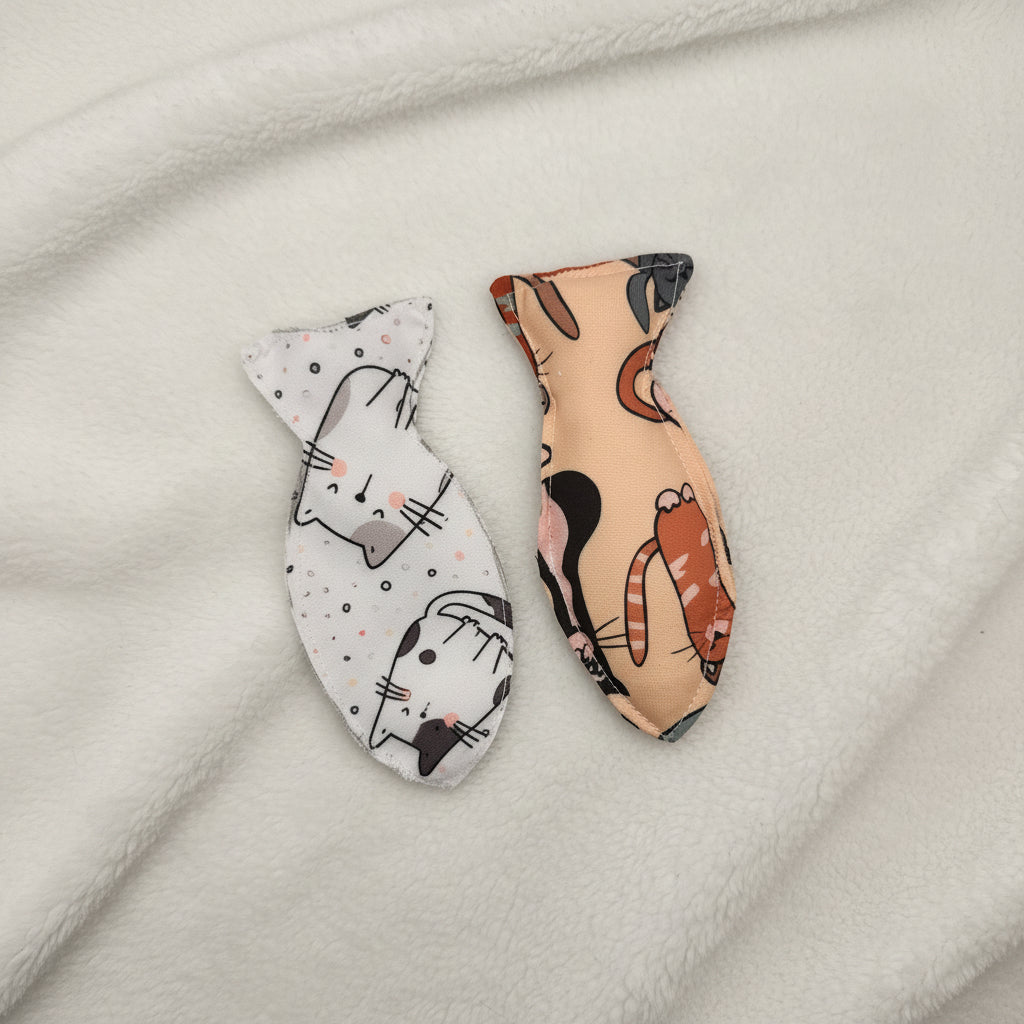 Two small fish-shaped fabric cat w with cat print patterns, one in beige with orange and black cat illustrations and the other in white with black and pink cat illustrations, displayed on a plain white surface.