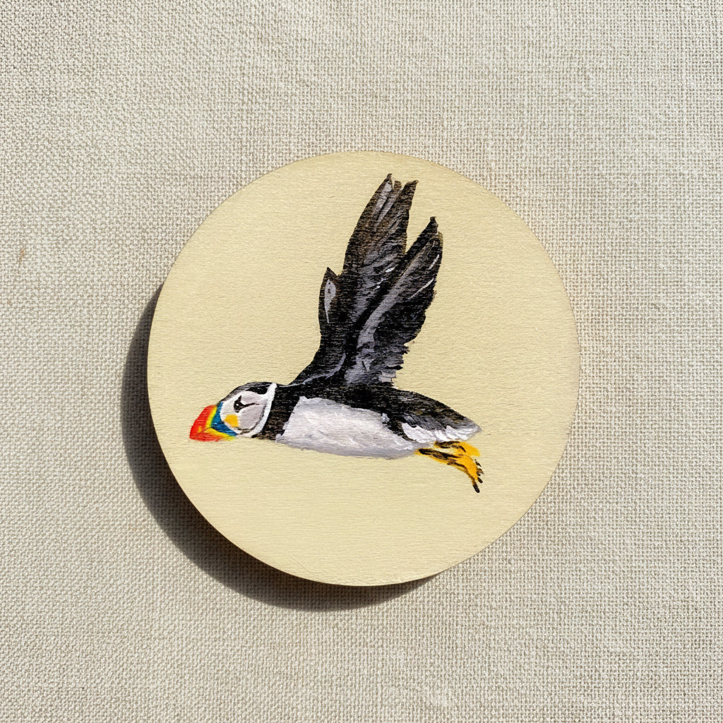 Hand-painted wooden disc with a depiction of a puffin flying against a beige background.