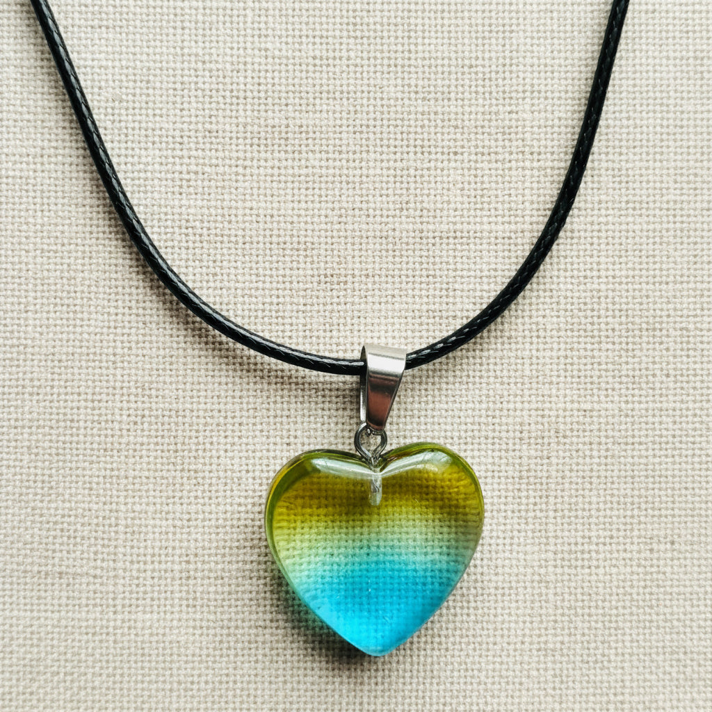 Heart-shaped blue-yellow pendant on a black cord against a neutral background