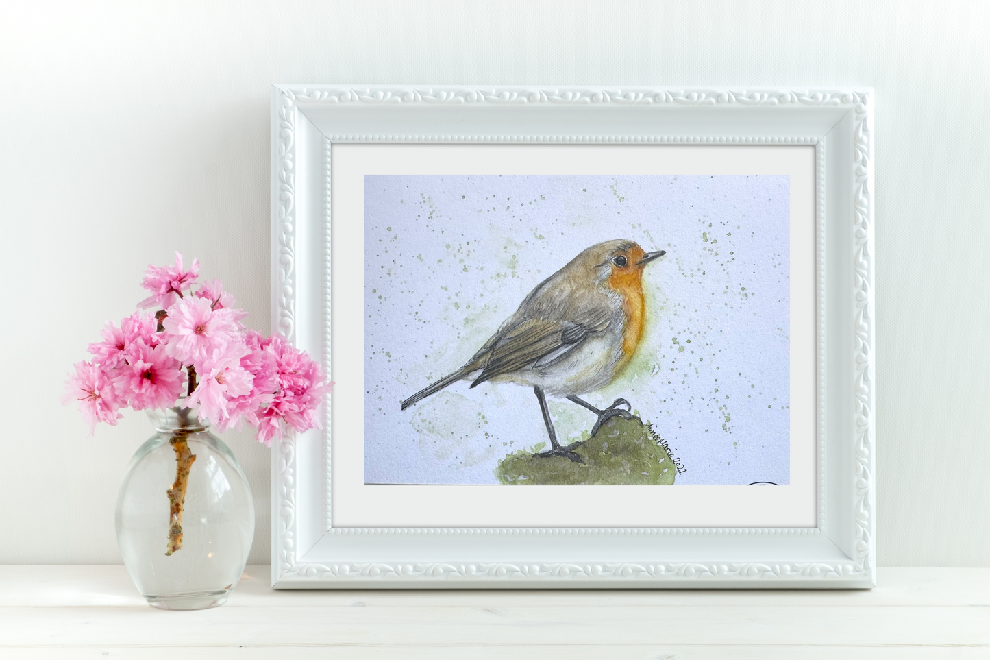 A watercolour print of a robin perched on a branch, framed and displayed on a white wall above a vase with pink flowers.