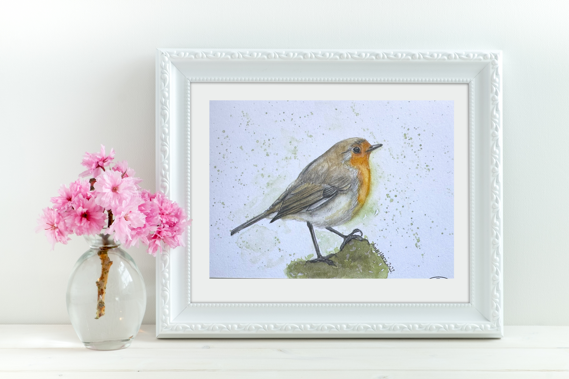 A watercolour print of a robin perched on a branch, framed and displayed on a white wall above a vase with pink flowers.