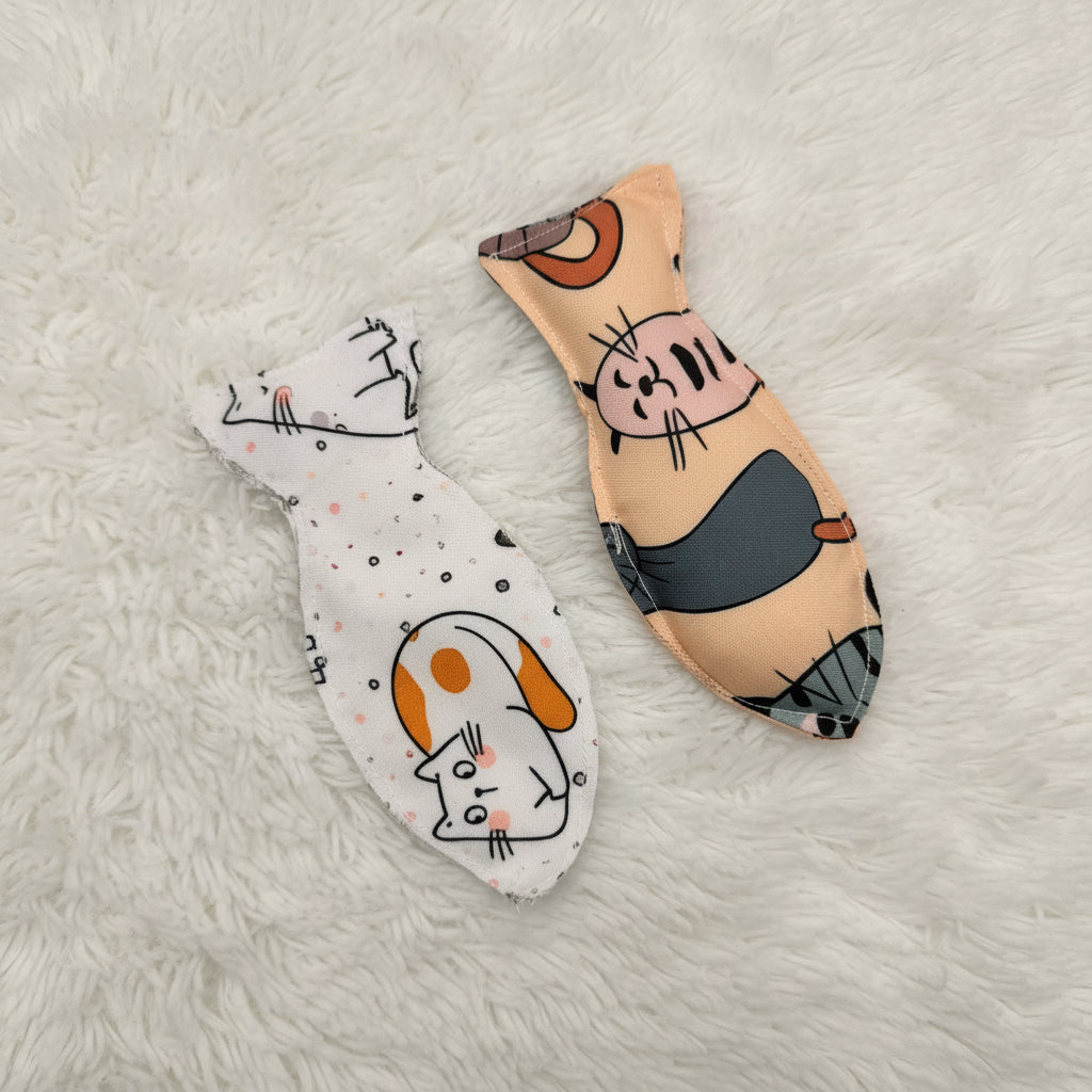 Two handmade fish-shaped fabric cat toys with cat face patterns, one in beige and the other in white, displayed on a plain light background.