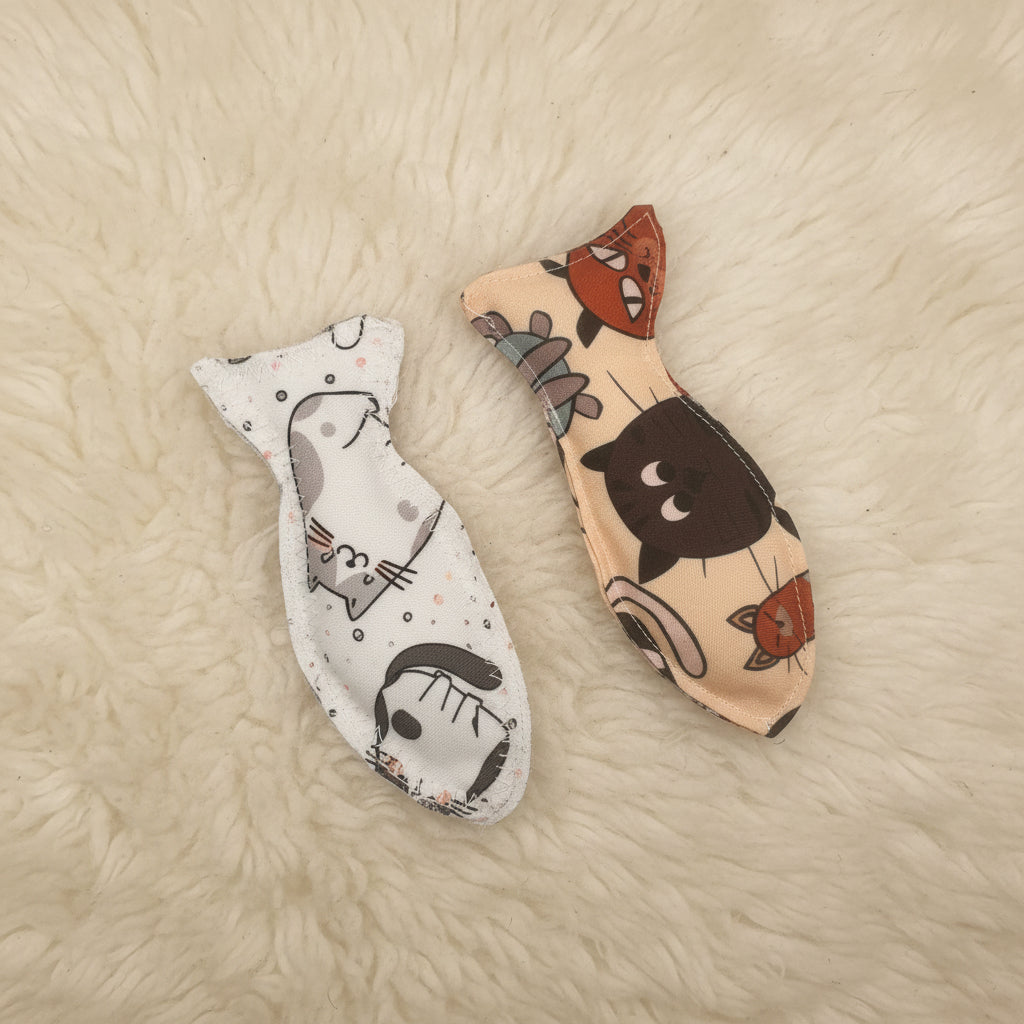 Two fish-shaped fabric cat toys on a plain light background. The top one has a peach background with a black cat face and small cat motifs.