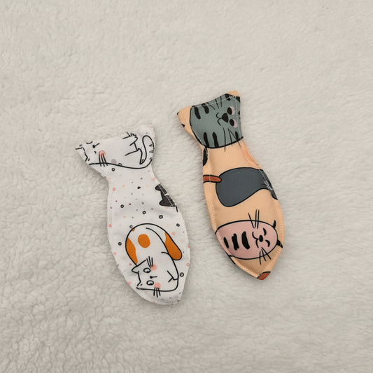 Two small fabric cat toys with printed cartoon cat faces, placed on a plain light background. One is white with black line art and orange accents, and the other is peach with gray and pink cats