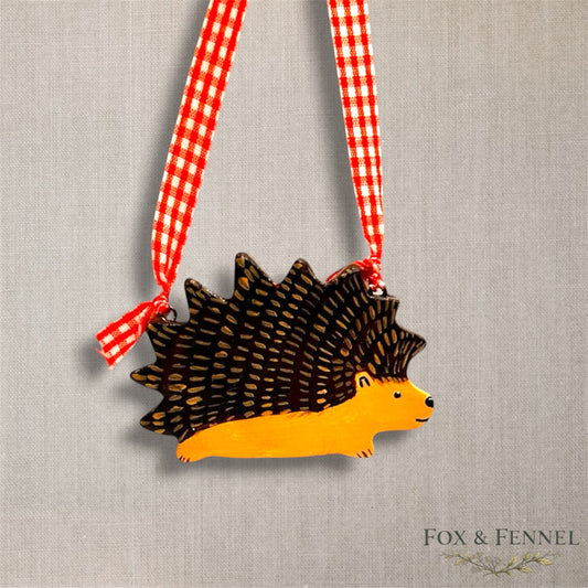 Clay Hedgehog hanging ornament with a red and white checkered ribbon on a textured gray background, branded 'FOX & FENNEL'.