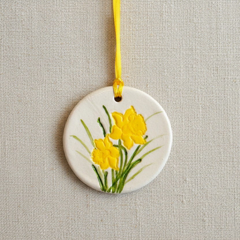 Round clay hanging ornament with yellow daffodil flowers on a pale beige background, yellow ribbon to hang