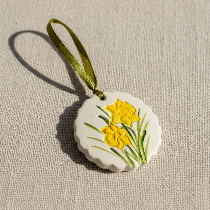 Decorative circular clay hanging ornament scalloped edges with yellow daffodil flowers on a pale beige background, green ribbon to hang