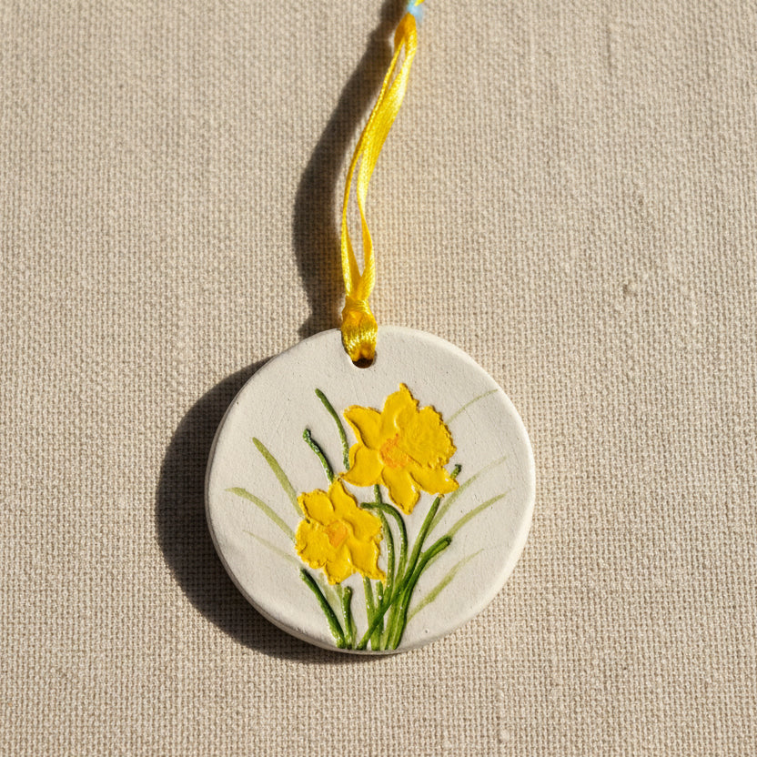 Round clay hanging ornament with yellow daffodil flowers on a pale beige background, yellow ribbon to hang
