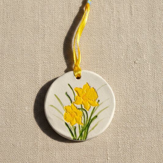 Round clay hanging ornament with yellow daffodil flowers on a pale beige background, yellow ribbon to hang