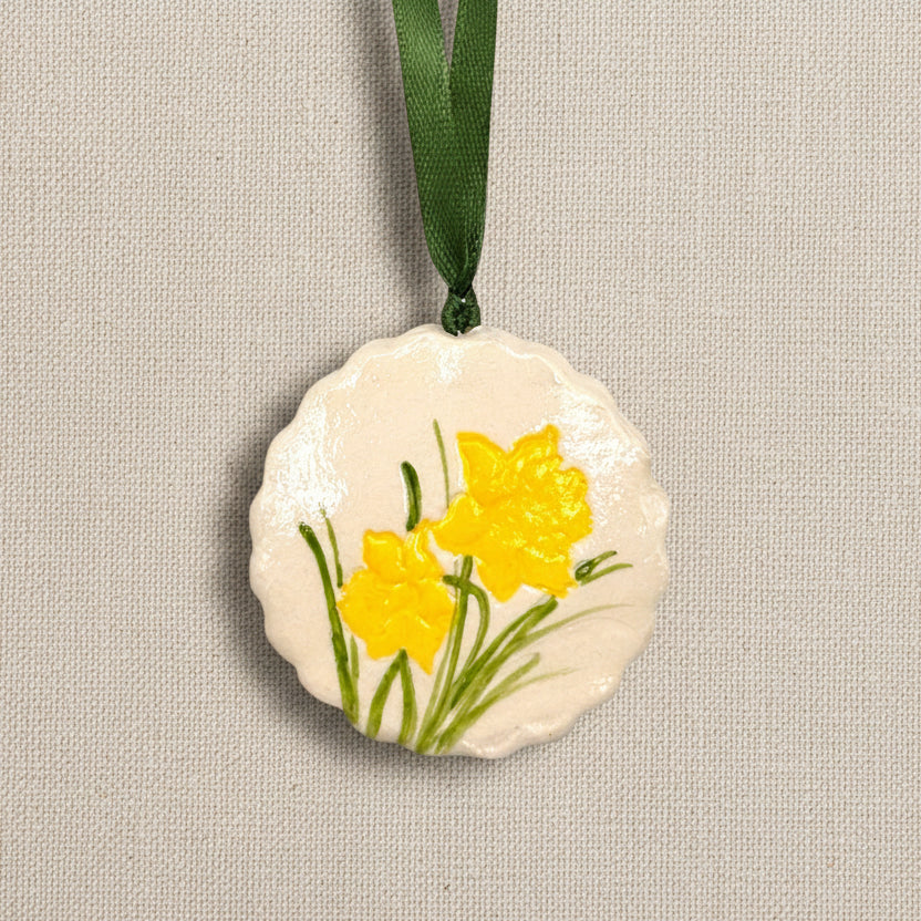 Decorative circular clay ornament with scalloped edge and yellow daffodil flowers on a light beige background, dark green ribbon to hang