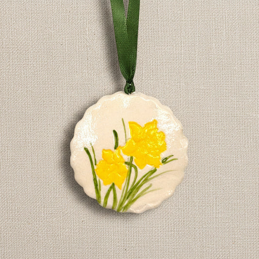 Decorative circular clay ornament with scalloped edge and yellow daffodil flowers on a light beige background, dark green ribbon to hang