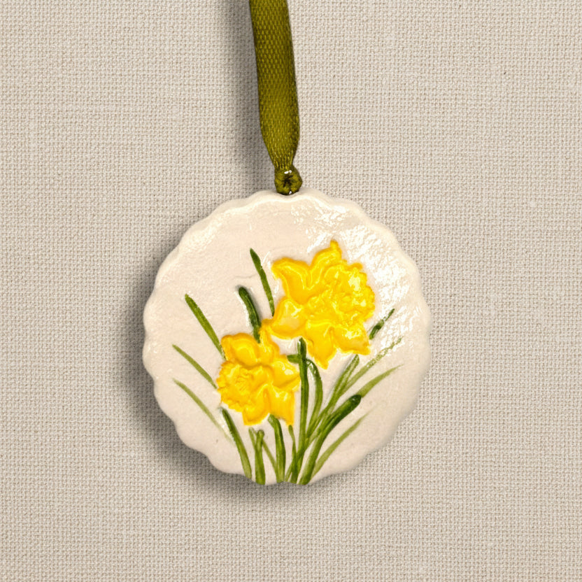 Decorative circular clay hanging ornament with scalloped edge with yellow daffodil flowers on a pale beige background, green ribbon to hang