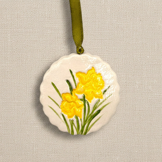 Decorative circular clay hanging ornament with scalloped edge with yellow daffodil flowers on a pale beige background, green ribbon to hang