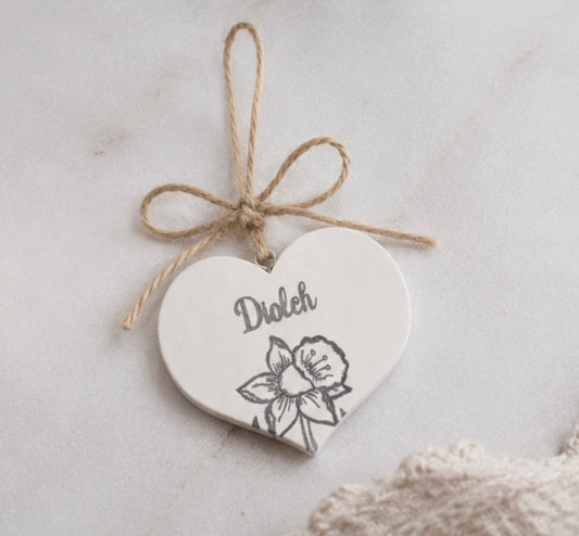 A white heart-shaped wooden ornament tied with a natural jute twine bow, featuring the word 'Dioch', Welsh for thank you, and a black line-art daffodil illustration, set against a light neutral background.