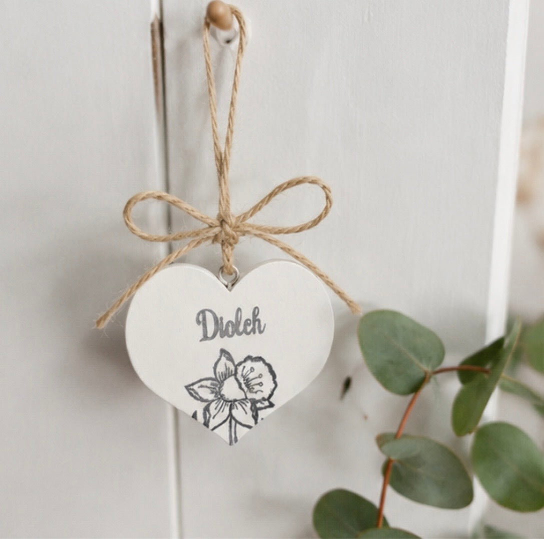 A white heart-shaped hanging ornament with a natural jute twine bow and loop, stamped with the word ‘Diolch' and a daffodil illustration, hanging on a white paneled door with eucalyptus leaves in the foreground.