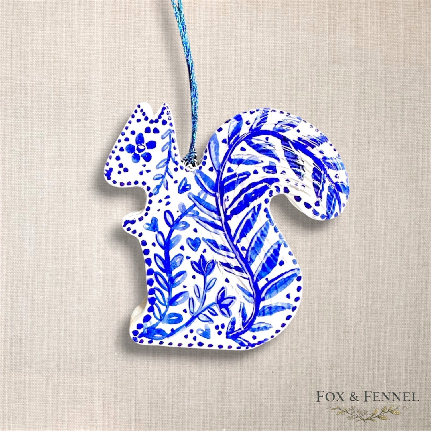 Decorative squirrel clay hanging ornament with blue floral patterns on an off white background, blue string to hang,  branded 'FOX & FENNEL'.