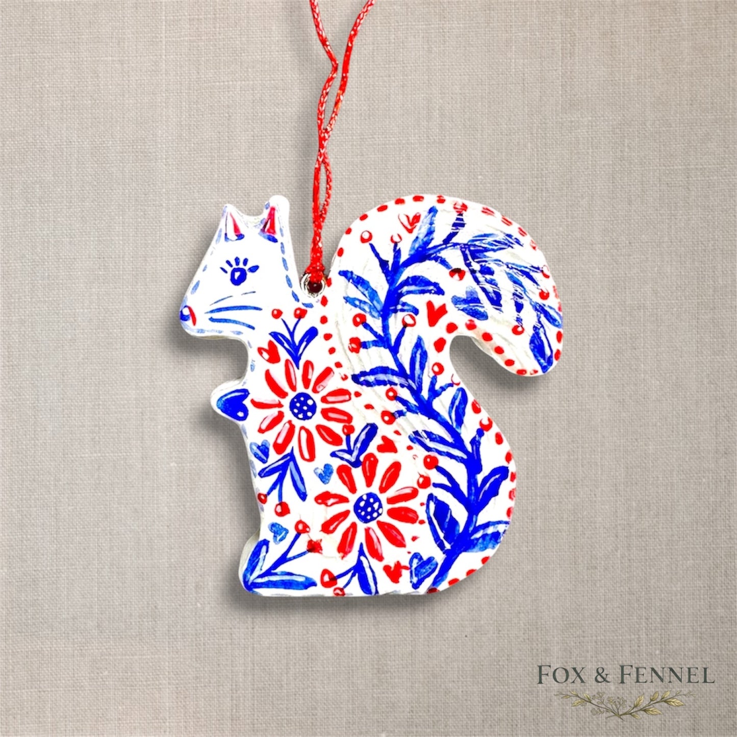 Decorative clay squirrel ornament with floral patterns in red, white, and blue on a beige fabric background. Red string to hang 