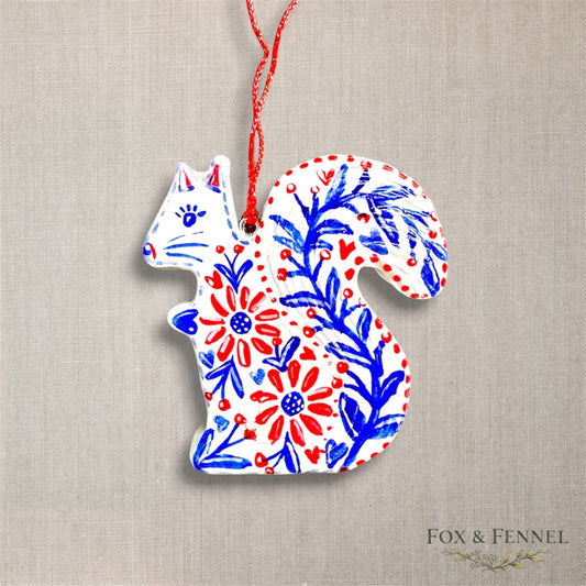 Decorative clay squirrel ornament with floral patterns in red, white, and blue on a beige fabric background. Red string to hang 