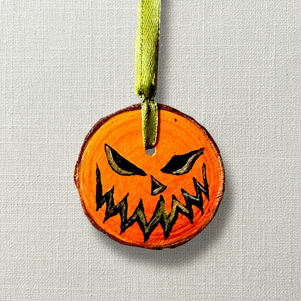 A hand-painted, natural wood slice decoration in the shape of a pumpkin with a green ribbon for hanging.