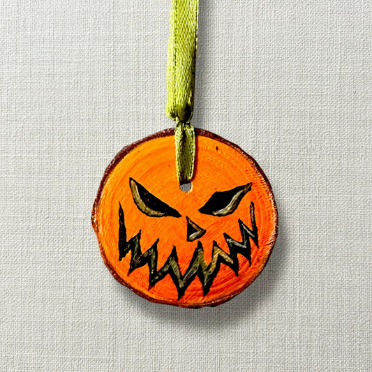 A hand-painted, natural wood slice decoration in the shape of a pumpkin with a green ribbon for hanging.