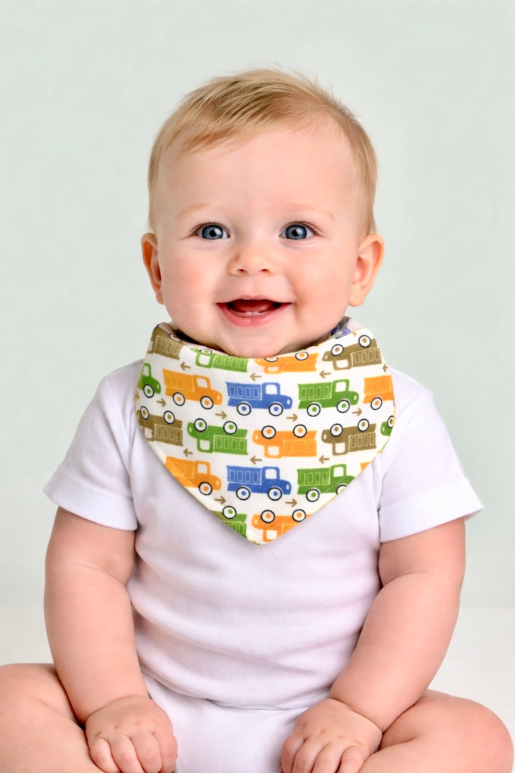 Baby wearing a white shirt with a colorful truck-patterned bib against a plain background