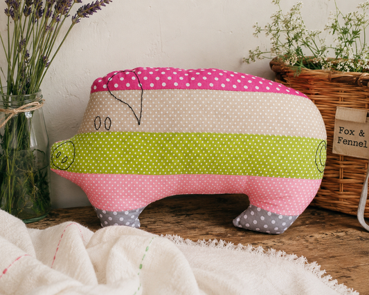 A handmade fabric pig-shaped with horizontal stripes of pink, beige, and lime green polka dot patterns, styled on a rustic wooden table with dried lavender and a wicker basket in the background.