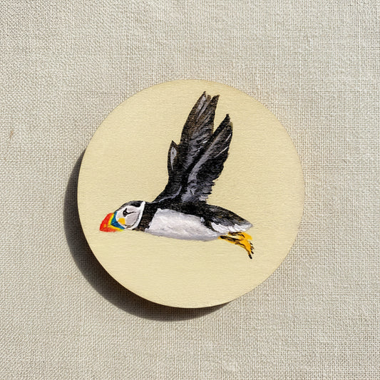 Hand-painted wooden disc with a depiction of a puffin flying against a beige background.