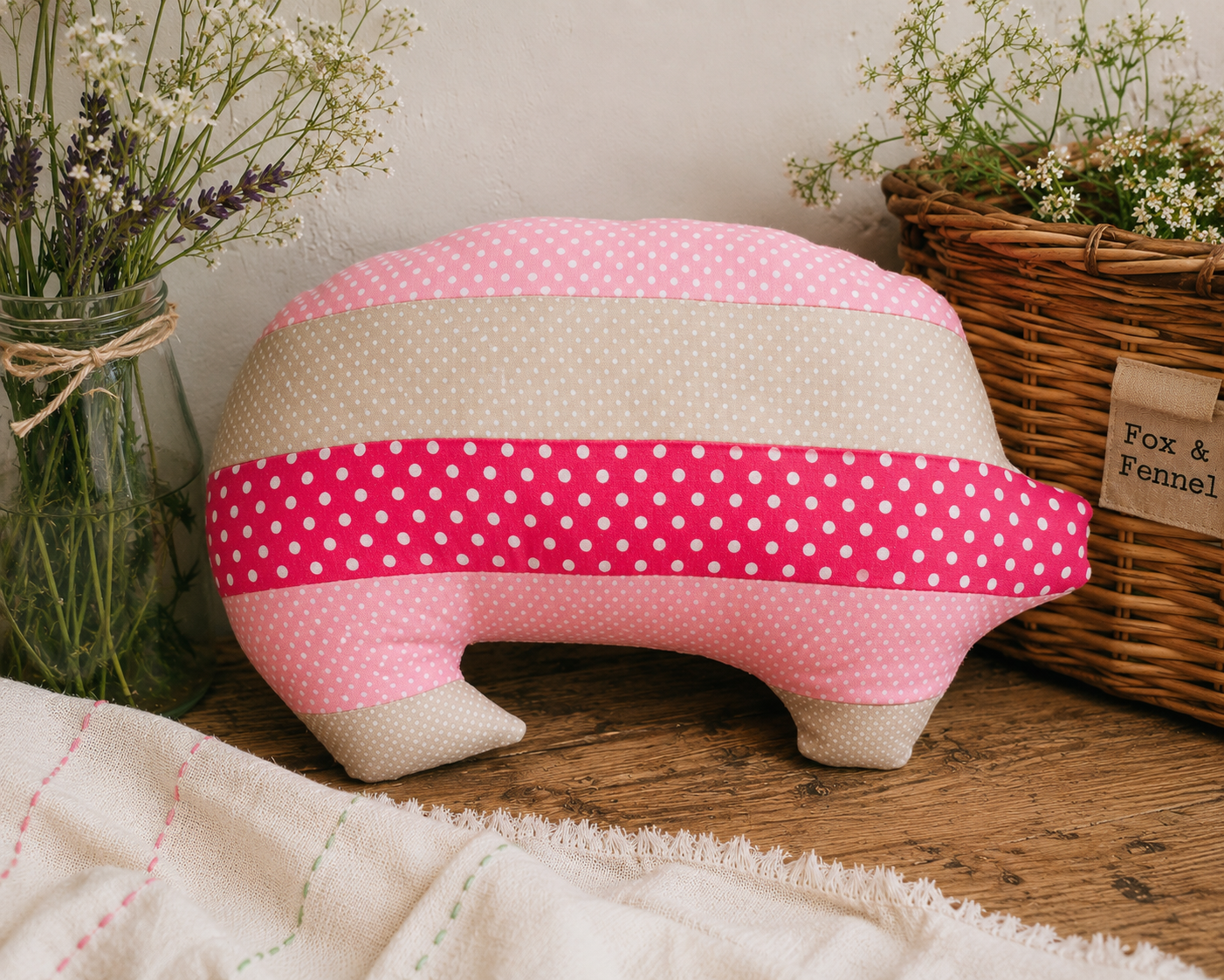 A handmade fabric pig-shaped cushion with pink and beige polka dot stripes, styled on a rustic wooden table with dried flowers and a wicker basket in the background.