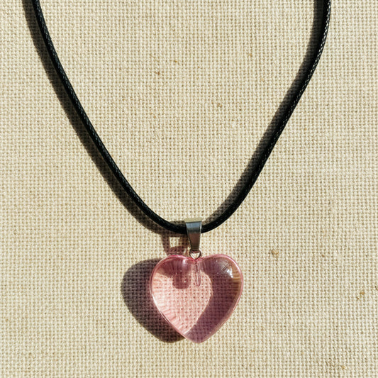 Heart-shaped pink transparent pendant on a black cord against a neutral background