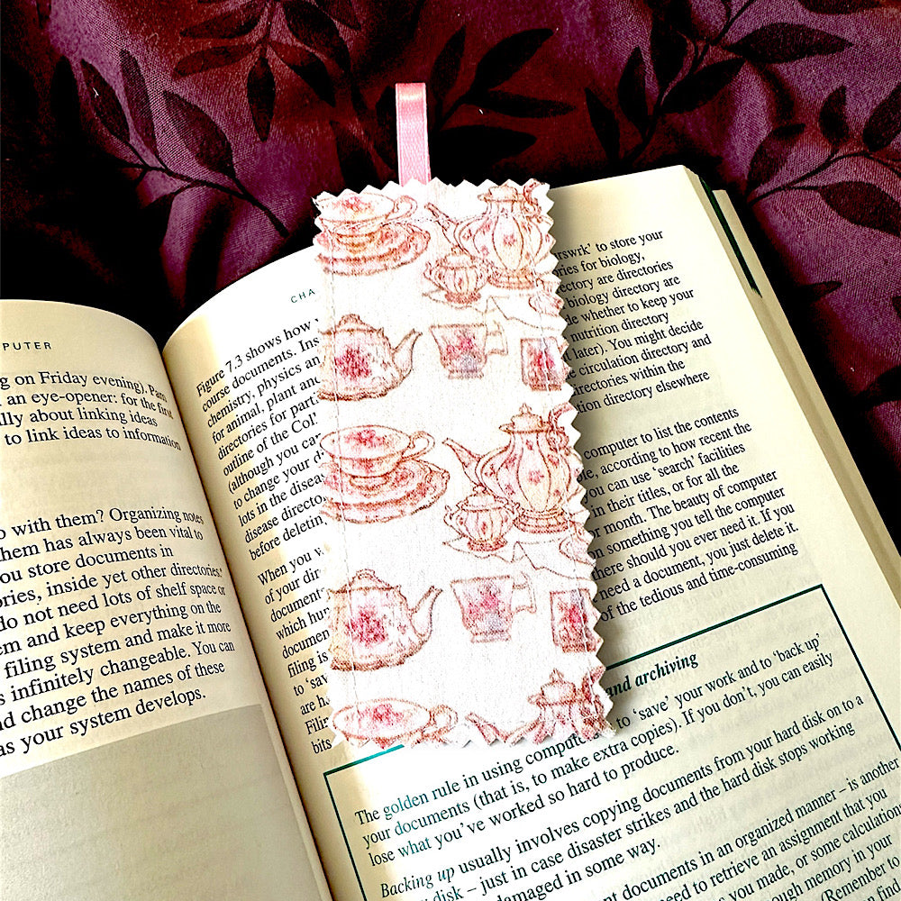 A handmade fabric bookmark with a pink teapot and teacups design, placed inside an open book.