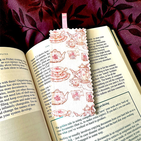 A handmade fabric bookmark with a pink teapot and teacups design, placed inside an open book.