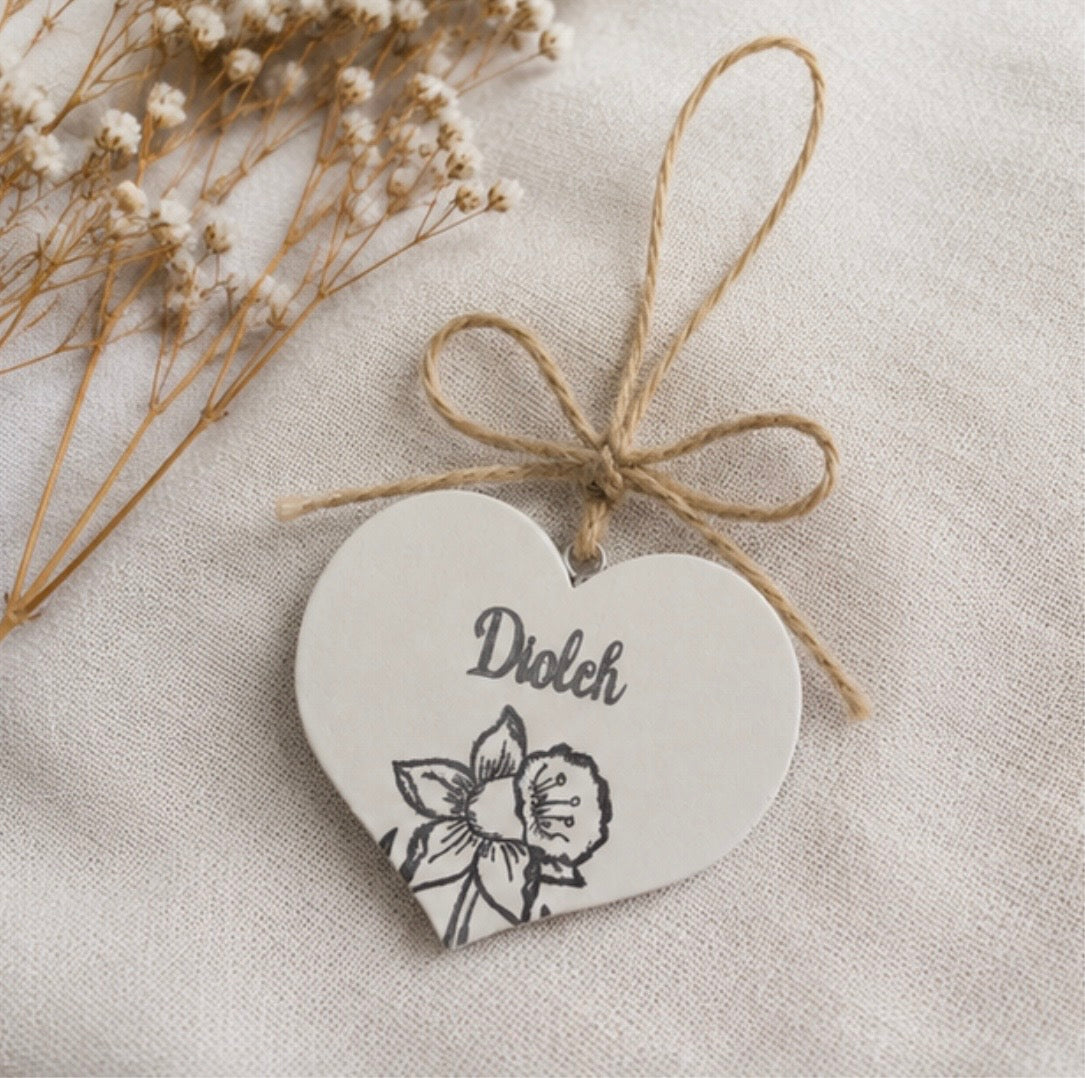 A heart-shaped white wooden hanging ornament tied with natural jute twine, featuring the word ‘Diolch', Welsh for thank you, and a black stamped daffodil, styled on a textured cream fabric with dried baby's breath.
