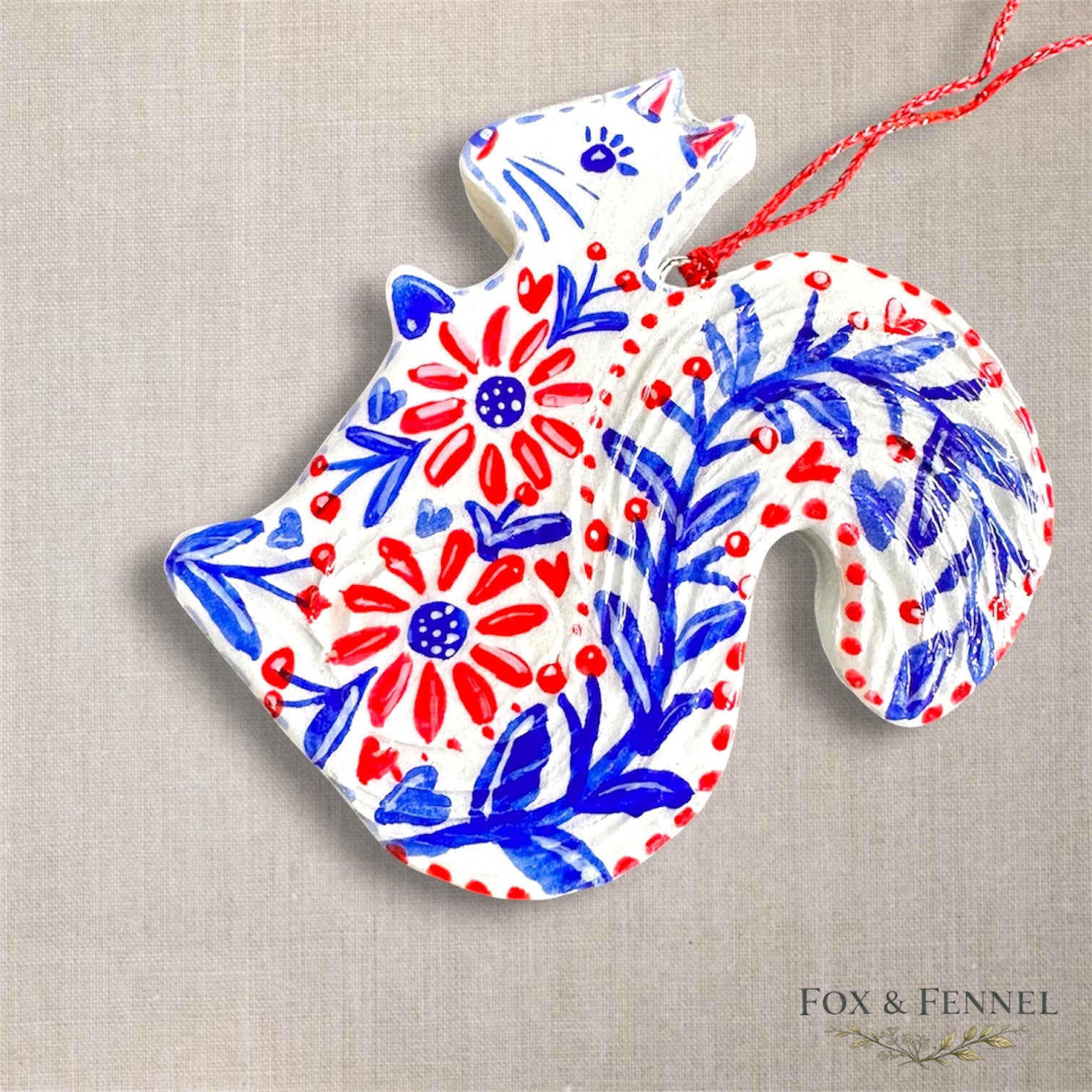 Decorative clay ornament shaped like a squirrel with floral patterns in red,white and blue. Red string to hang 