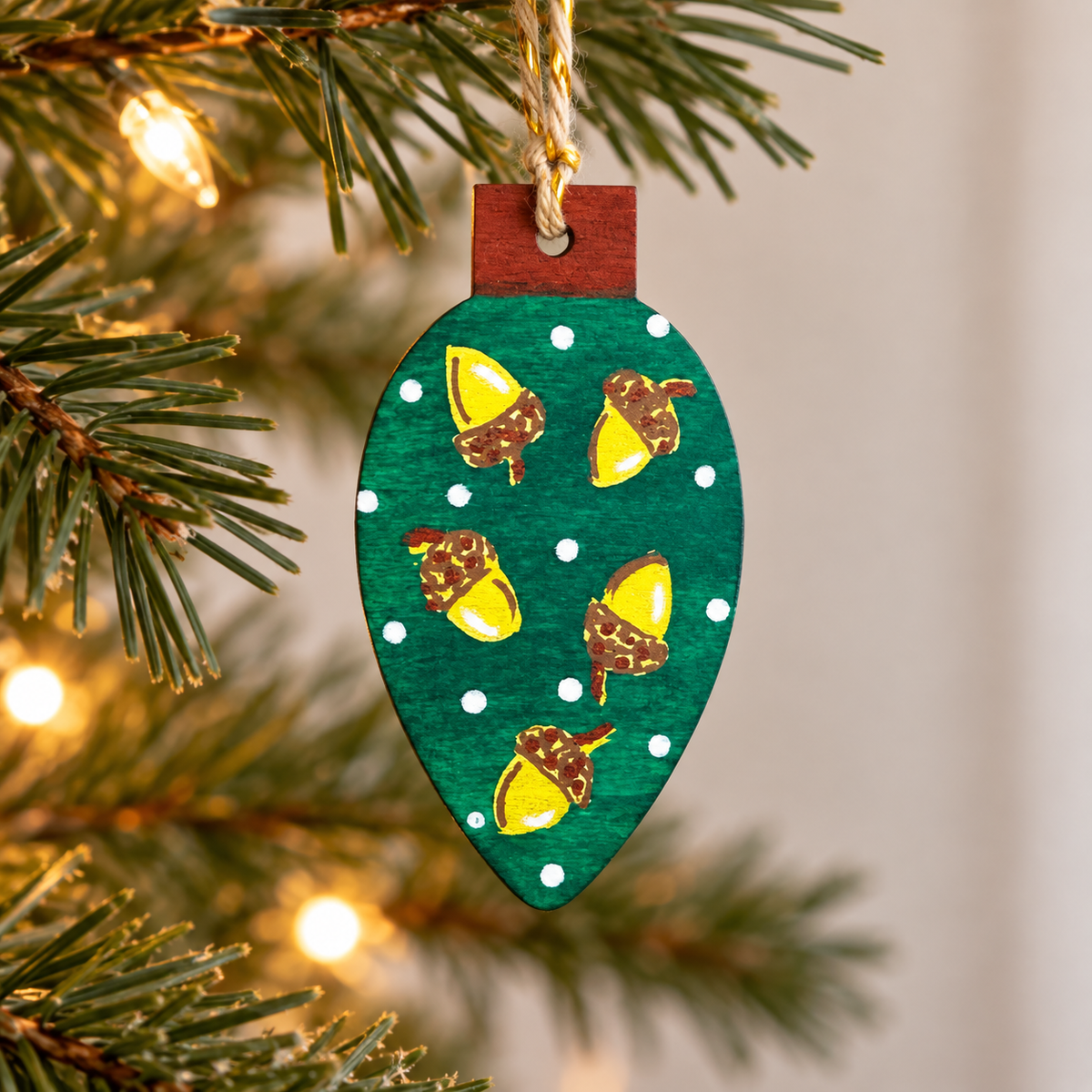 Hand-painted green wooden Christmas ornament decorated with golden acorns and white dots, hanging from a twisted cord on a Christmas tree
