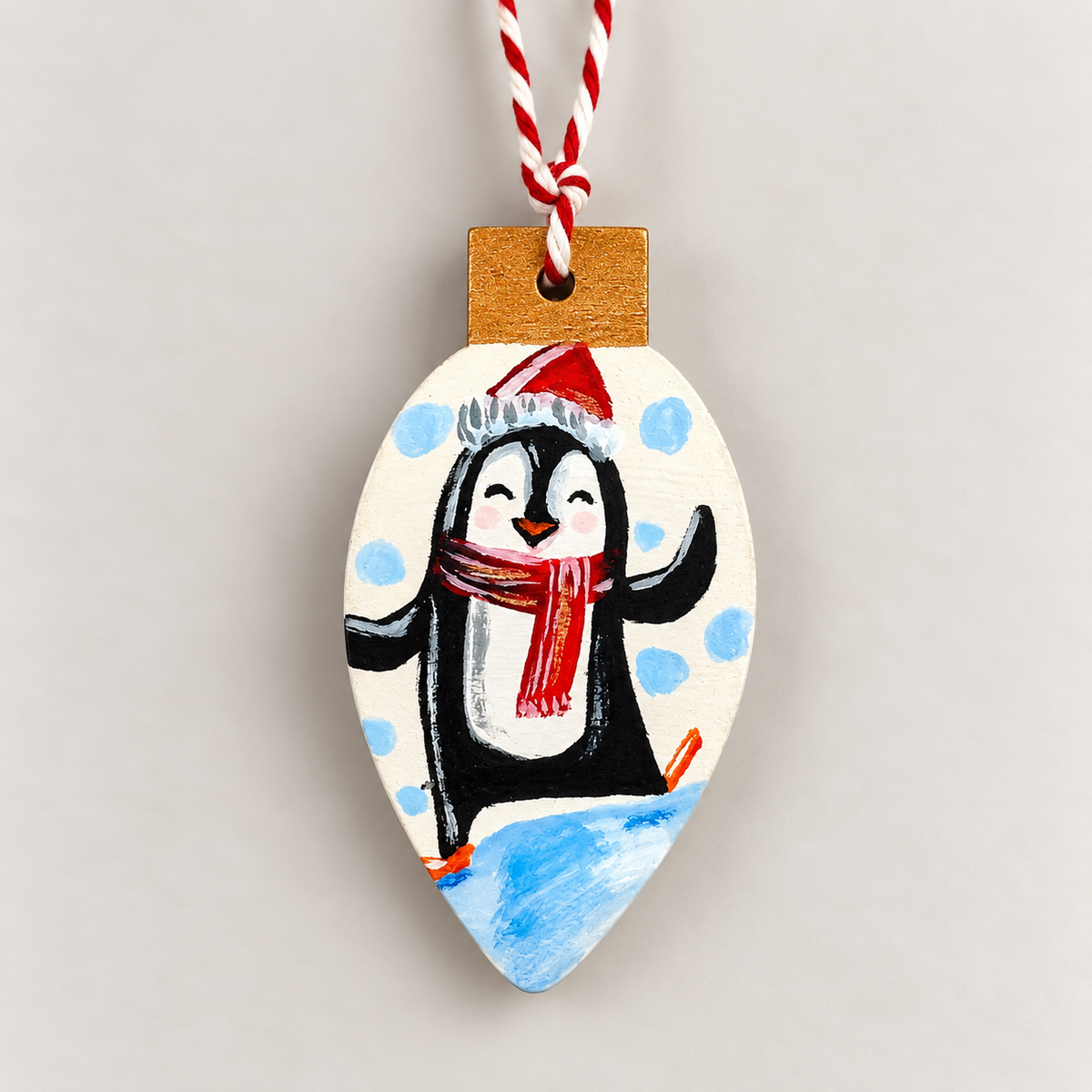A hand-painted wooden Christmas ornament shaped like a light bulb, featuring a penguin wearing a red Santa hat and scarf on a white background with blue polka dots and a blue snowy base. It has a red and white striped hanging string and a natural wood cap.