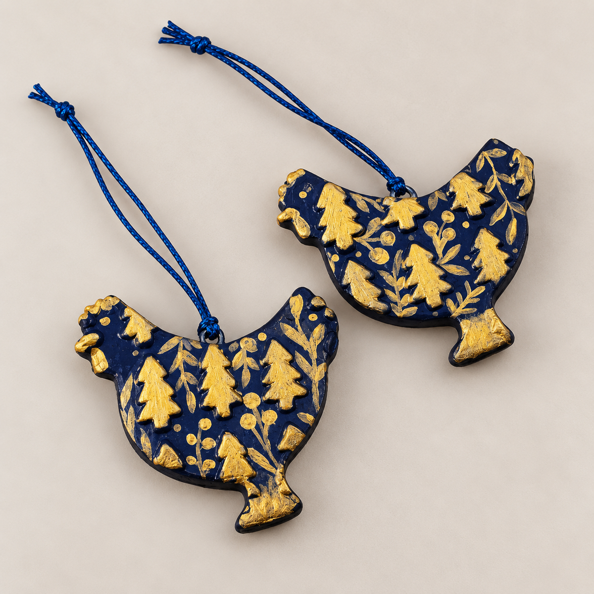 Two handmade dark blue clay chicken Christmas ornaments decorated with gold Christmas trees and botanical details