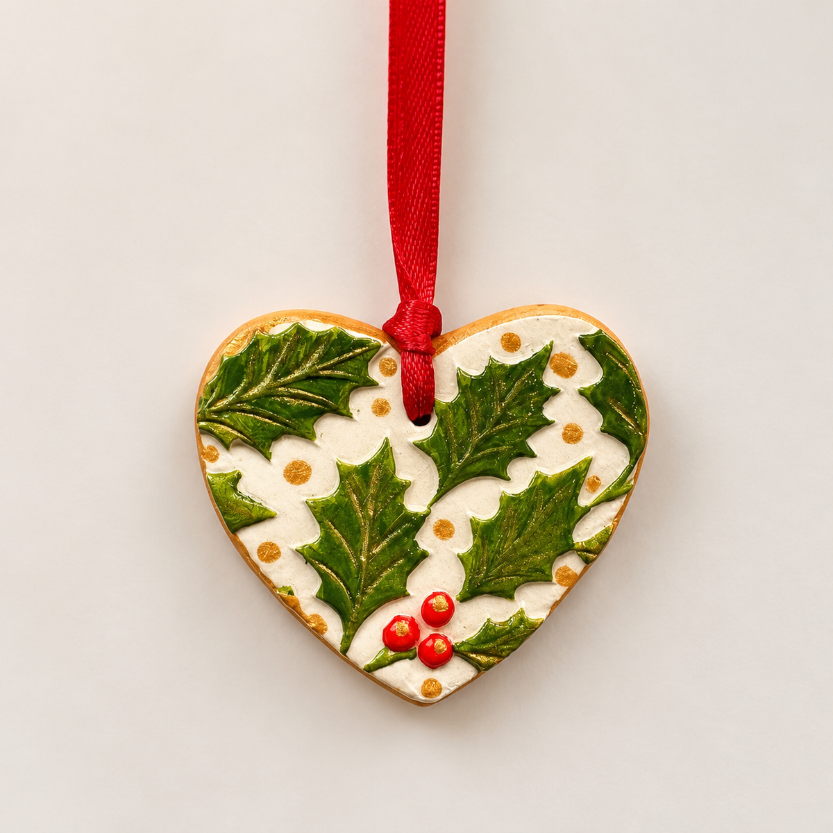 Handmade heart-shaped clay Christmas tree decoration with embossed green holly leaves, red berries, cream background, metallic gold detailing and a red hanging ribbon.