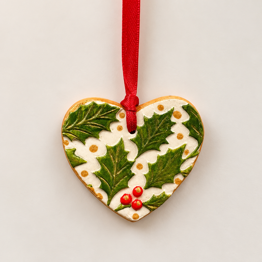 Handmade heart-shaped clay Christmas tree decoration with embossed green holly leaves, red berries, cream background, metallic gold detailing and a red hanging ribbon.