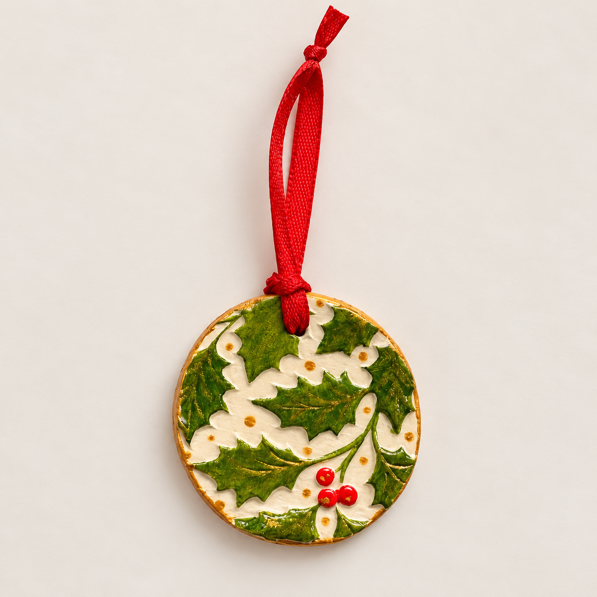 Round handmade clay Christmas tree decoration with embossed green holly leaves, three red berries, cream background, metallic gold detailing and a red hanging ribbon.