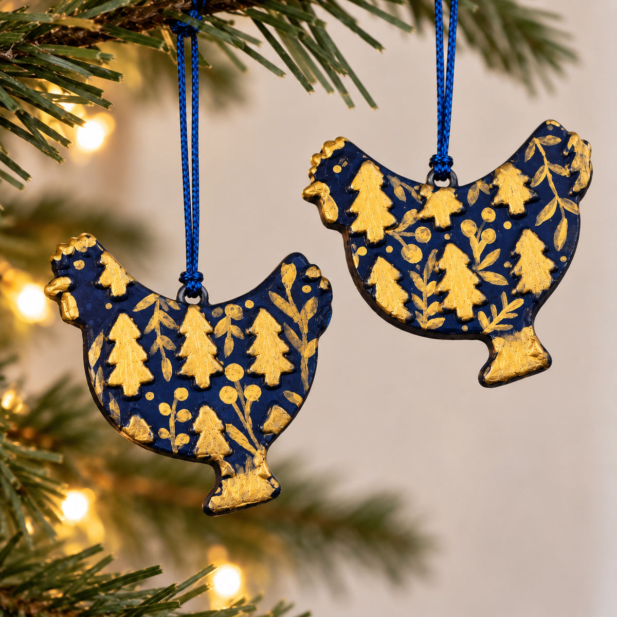 Two handmade dark blue clay chicken Christmas ornaments decorated with gold Christmas trees and botanical details