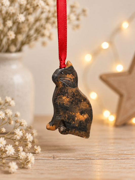Handmade tortoiseshell cat clay Christmas ornament with a red ribbon, photographed against a warm neutral background with soft fairy lights.
