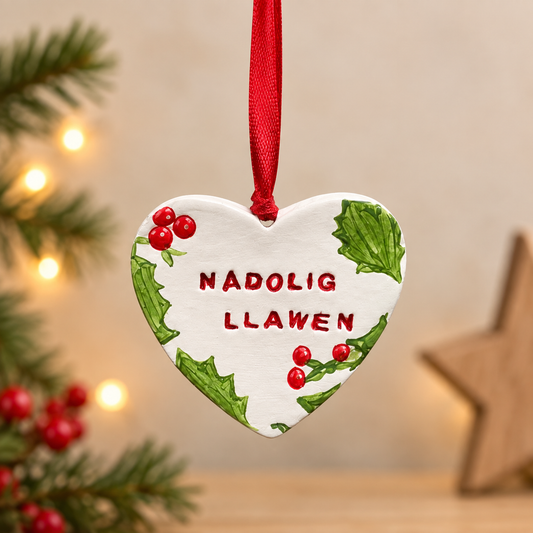 Handmade clay Christmas heart ornament with hand-painted holly, red berries and “Nadolig Llawen” lettering, finished with a red ribbon.