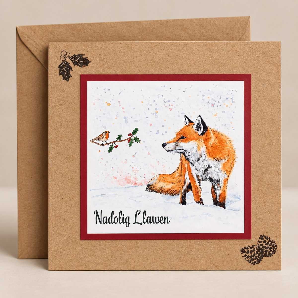15 x 15 cm natural Kraft Welsh Christmas card featuring a watercolour fox and robin in a snowy winter scene with holly and the words Nadolig Llawen